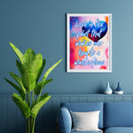&quot;Love is the paint that makes our hearts a masterpiece&quot; fine art giclee print, with a 1/2&quot; white mat, in a 1/2&quot; white picture frame, hanging on teal blusish wall over a blue couch and plant.